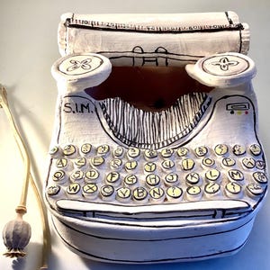 Peut inclure: Sculpture de machine à écrire en céramique blanche avec des contours noirs et des touches jaunes. La sculpture comporte un rouleau de papier en haut et deux touches rondes de chaque côté. Deux capsules de graines séchées sont au premier plan.