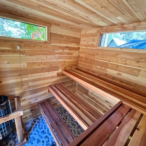 May include: A sauna interior featuring horizontal cedar wood panelling on the walls and ceiling, two tiered wooden benches, and a metal heater unit. Two rectangular windows are positioned high on the walls, and the floor is covered in small blue stones.