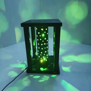 Può includere: Una lanterna rettangolare verde scuro con un nucleo cilindrico perforato. Il nucleo è illuminato da piccole luci brillanti. La lanterna ha una base e una parte superiore quadrate, con supporti verticali. Un cavo di alimentazione nero si estende dalla base.