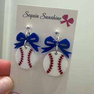 May include: Baseball-themed earrings featuring white circular pendants with red stitching, topped with a blue bow. The earrings are attached to a silver post. The card behind the earrings says "Sequin Sunshine" and has a website address.