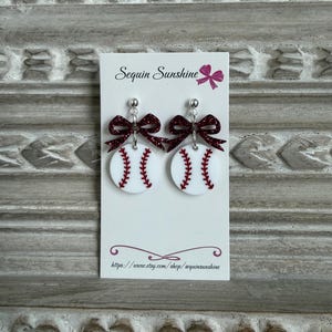 May include: Baseball-themed earrings featuring white circular pendants with red stitching, mimicking baseballs. Each earring has a sparkly burgundy bow and a silver stud. The earrings are displayed on a white card with the text "Sequin Sunshine".