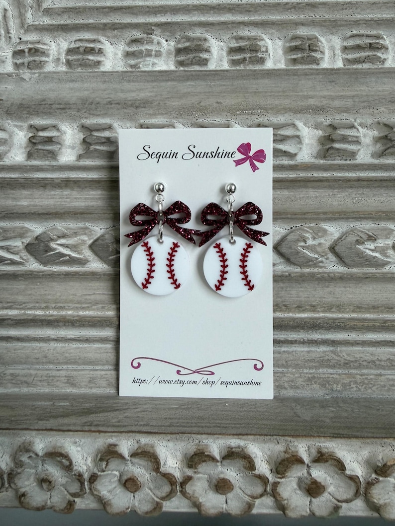 May include: Baseball-themed earrings featuring white circular pendants with red stitching, mimicking baseballs. Each earring has a sparkly burgundy bow and a silver stud. The earrings are displayed on a white card with the text "Sequin Sunshine".