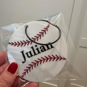 Pu&ograve; includere: Portachiavi rotondo bianco a tema baseball con cuciture rosse e il nome "Julian" in lettere nere.