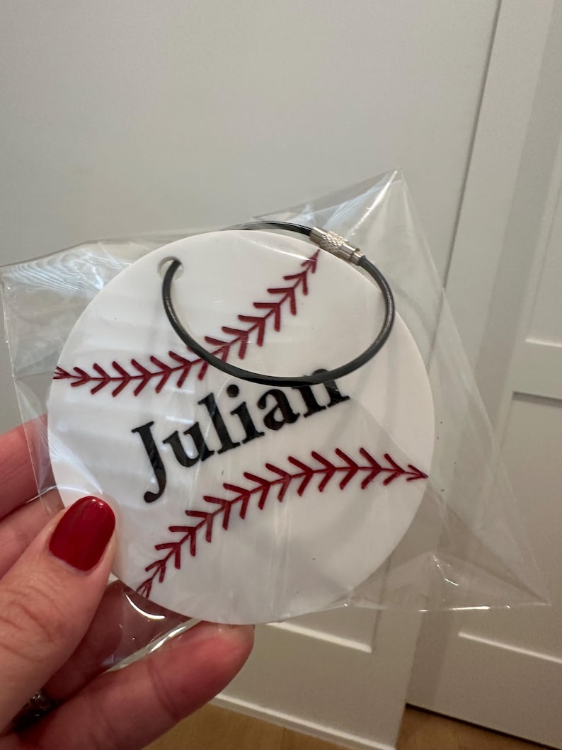 Pu&ograve; includere: Portachiavi rotondo bianco a tema baseball con cuciture rosse e il nome "Julian" in lettere nere.