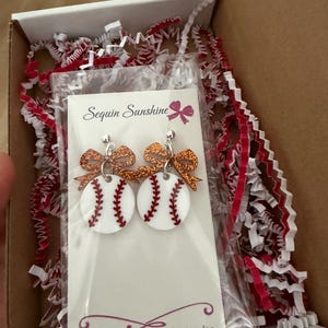 May include: Baseball-themed earrings with white circles, red stitching, and glittery orange bows. The earrings are packaged in a box with red and white crinkle paper. The card reads "Sequin Sunshine" and the Etsy shop URL.