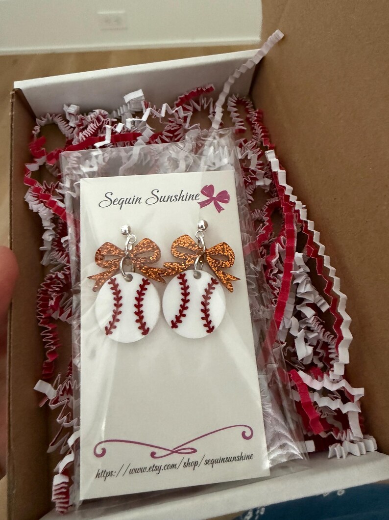 May include: Baseball-themed earrings with white circles, red stitching, and glittery orange bows. The earrings are packaged in a box with red and white crinkle paper. The card reads "Sequin Sunshine" and the Etsy shop URL.
