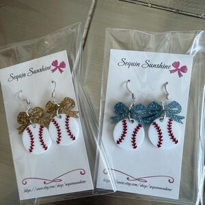 May include: Baseball-themed earrings with white circular bases, red stitching, and glittery bow accents. One pair has gold bows, the other has blue bows. Each pair is packaged on a white card with the text "Sequin Sunshine".