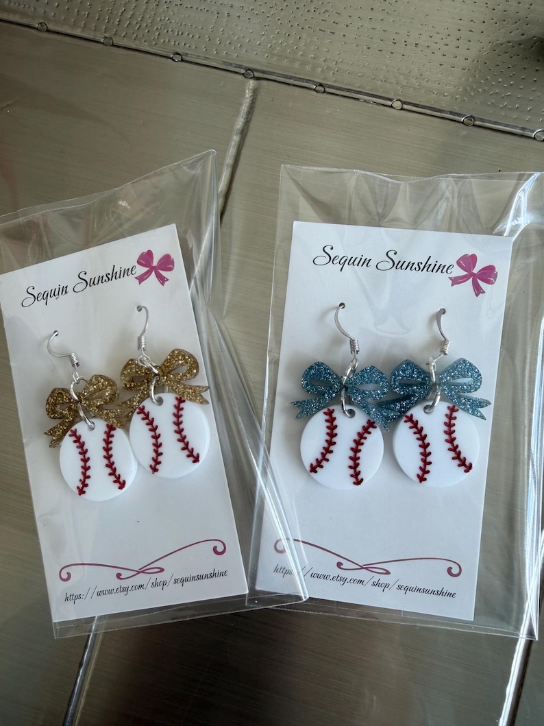 May include: Baseball-themed earrings with white circular bases, red stitching, and glittery bow accents. One pair has gold bows, the other has blue bows. Each pair is packaged on a white card with the text "Sequin Sunshine".