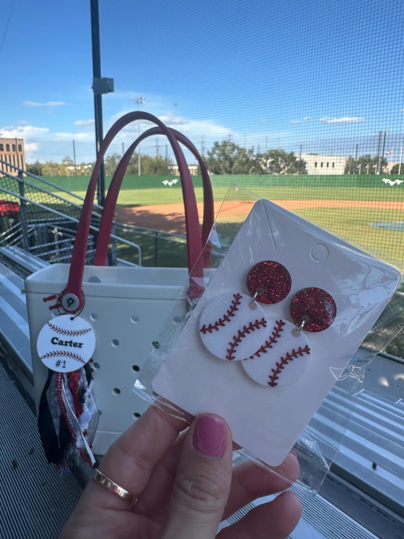 May include: White tote bag with red handles and a baseball-themed tag that says "Carter #1". Baseball earrings with red glitter tops and white baseball-shaped bottoms with red stitching are also visible.