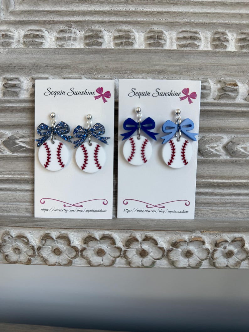May include: Baseball-themed earrings on white cards. The earrings feature a white circular design with red stitching, resembling a baseball. Each earring has a bow at the top, with one pair featuring a glittery navy bow and the other pair with a navy and light blue bow.