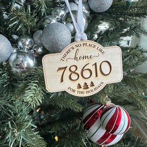 May include: A wooden Christmas ornament with the text "There's no place like home 78610 for the holidays." The ornament is hanging on a Christmas tree with silver ornaments and red and white striped ornaments.
