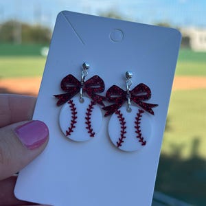 May include: Baseball-themed earrings featuring white circles with red stitching, mimicking baseballs. Each earring has a sparkly red bow at the top and a silver stud. The earrings are displayed on a white card.