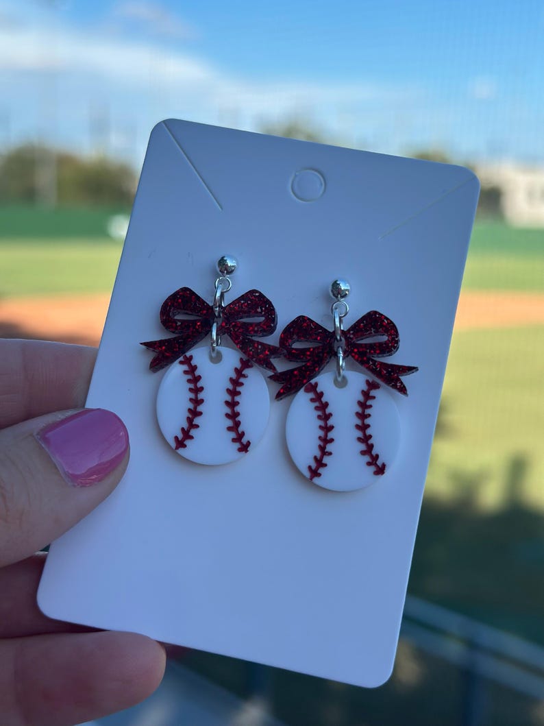May include: Baseball-themed earrings featuring white circles with red stitching, mimicking baseballs. Each earring has a sparkly red bow at the top and a silver stud. The earrings are displayed on a white card.