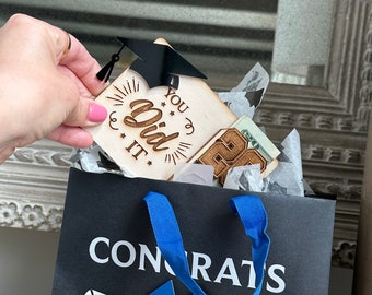 May include: A black gift bag with a white "Congrats" inscription and a blue ribbon tied around a white diploma. The bag is filled with tissue paper and a wooden sign that says "You Did It" with a black graduation cap on top.