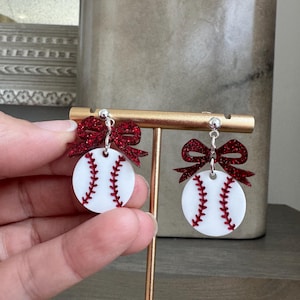 May include: Baseball-themed earrings. Each earring features a white circular design with red stitching, resembling a baseball. A sparkly red bow is attached above each baseball design. The earrings are displayed on a gold-colored stand.