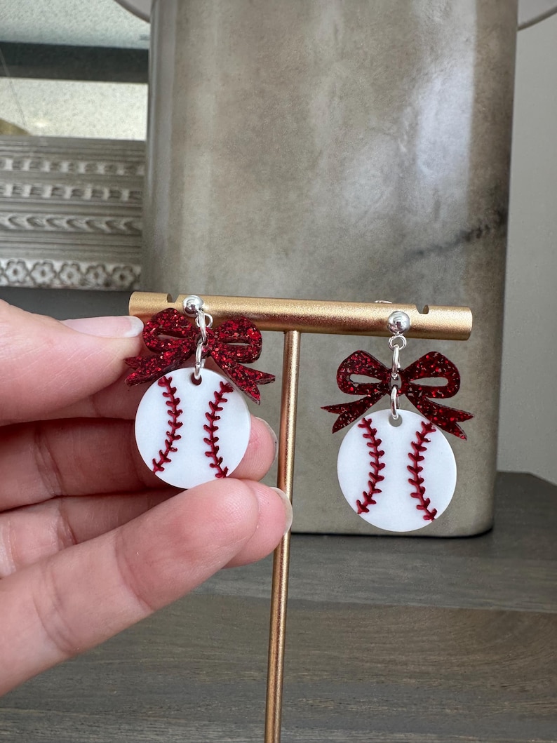 May include: Baseball-themed earrings. Each earring features a white circular design with red stitching, resembling a baseball. A sparkly red bow is attached above each baseball design. The earrings are displayed on a gold-colored stand.