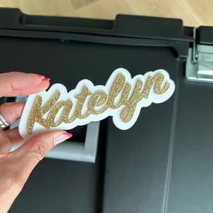 May include: A white, cursive nameplate with gold glitter lettering that reads "Katelyn".