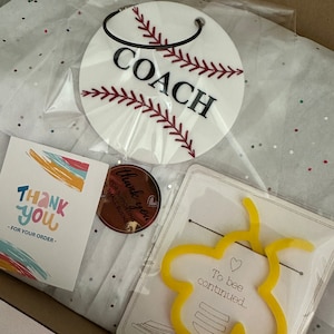 Pu&ograve; includere: Una scatola regalo piena di articoli, tra cui un portachiavi bianco a tema baseball con la parola "COACH" in lettere nere, un biglietto di ringraziamento con strisce colorate, un piccolo adesivo rotondo che dice "Grazie per aver sostenuto le piccole imprese", e un segnalibro giallo a forma di ape con il testo "To bee continued..." su un biglietto bianco.
