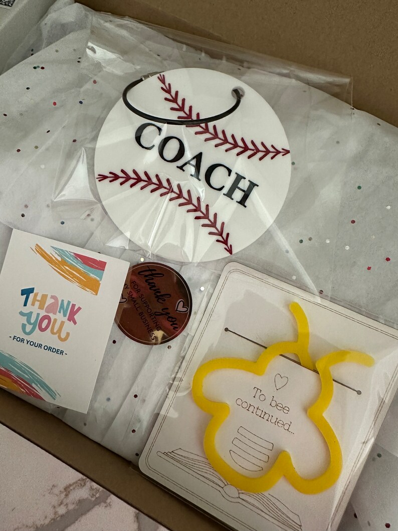 Pu&ograve; includere: Una scatola regalo piena di articoli, tra cui un portachiavi bianco a tema baseball con la parola "COACH" in lettere nere, un biglietto di ringraziamento con strisce colorate, un piccolo adesivo rotondo che dice "Grazie per aver sostenuto le piccole imprese", e un segnalibro giallo a forma di ape con il testo "To bee continued..." su un biglietto bianco.