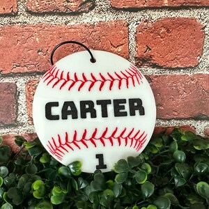Pu&ograve; includere: Ciondolo a forma di palla da baseball bianco con cuciture rosse e il nome "CARTER" in lettere nere. Il numero "1" &egrave; anche stampato in nero sull'ornamento.