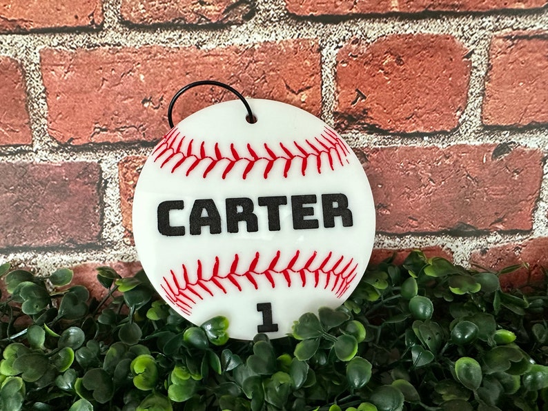 Pu&ograve; includere: Ciondolo a forma di palla da baseball bianco con cuciture rosse e il nome "CARTER" in lettere nere. Il numero "1" &egrave; anche stampato in nero sull'ornamento.