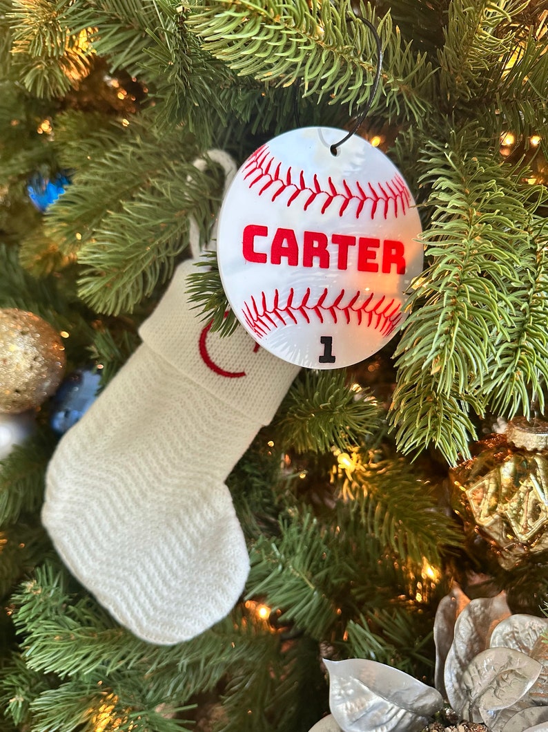 Pu&ograve; includere: Calza bianca a maglia con ricamo rosso appesa a un albero di Natale verde con un ornamento da baseball bianco con cuciture rosse e il nome "CARTER" e il numero "1" su di esso.