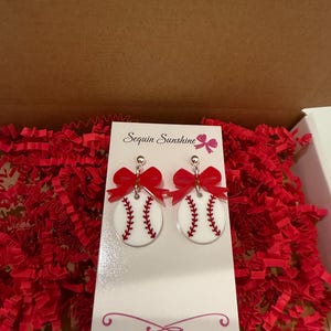 May include: White baseball earrings with red stitching and a red bow. The earrings are round and have a baseball design. The earrings are displayed on a white card with the text "Sequin Sunshine" and a website address.