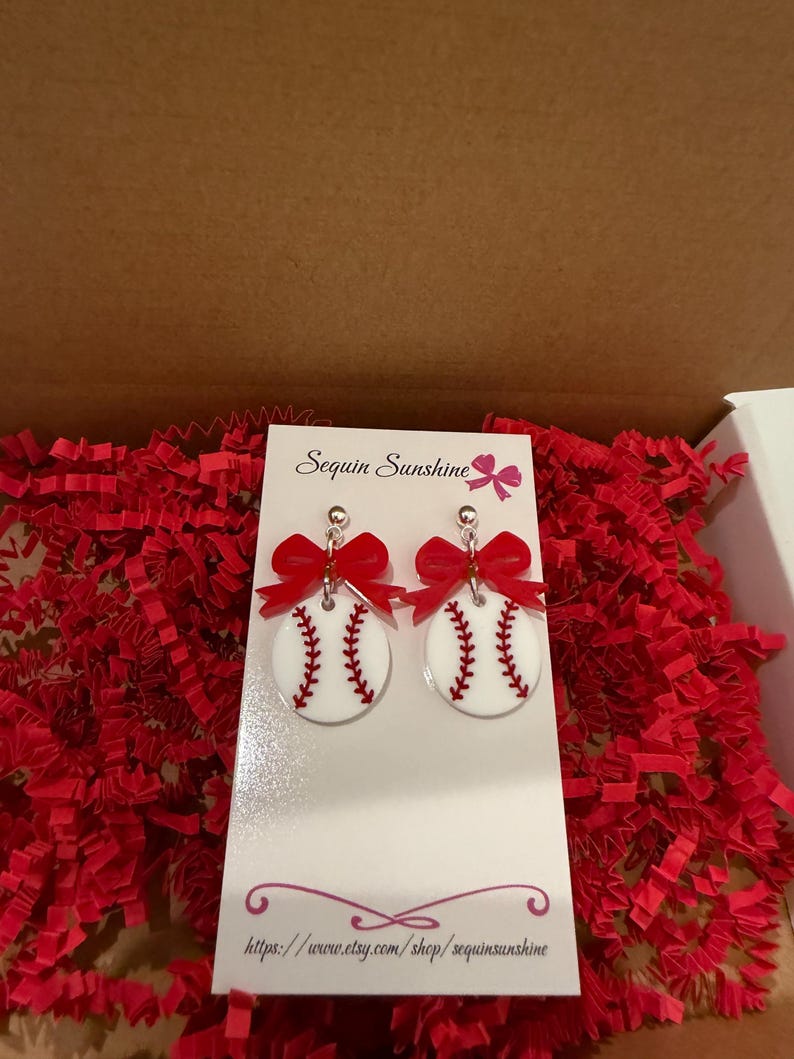May include: White baseball earrings with red stitching and a red bow. The earrings are round and have a baseball design. The earrings are displayed on a white card with the text "Sequin Sunshine" and a website address.