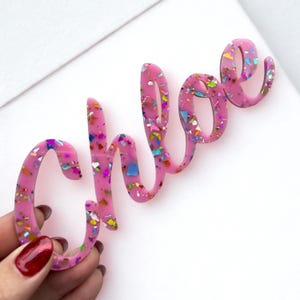 May include: A pink acrylic name sign featuring the name Chloe in a cursive script font. The material is translucent pink with embedded multicolored iridescent glitter flakes that reflect light in various shades of blue, green, and gold.