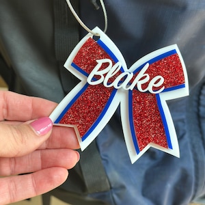 May include: A personalized luggage tag in the shape of a bow. The tag features the name "Blake" in white script over a red glitter background, with blue and white accents. It is attached to a silver wire loop.