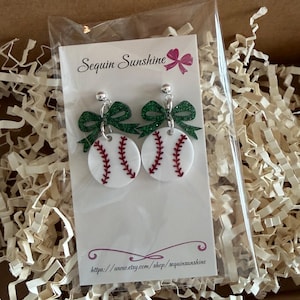 May include: Baseball-themed earrings with white baseballs featuring red stitching and green glitter bows. The earrings are displayed on a card with the shop name "Sequin Sunshine" and a website address, inside a cardboard box with paper filler.