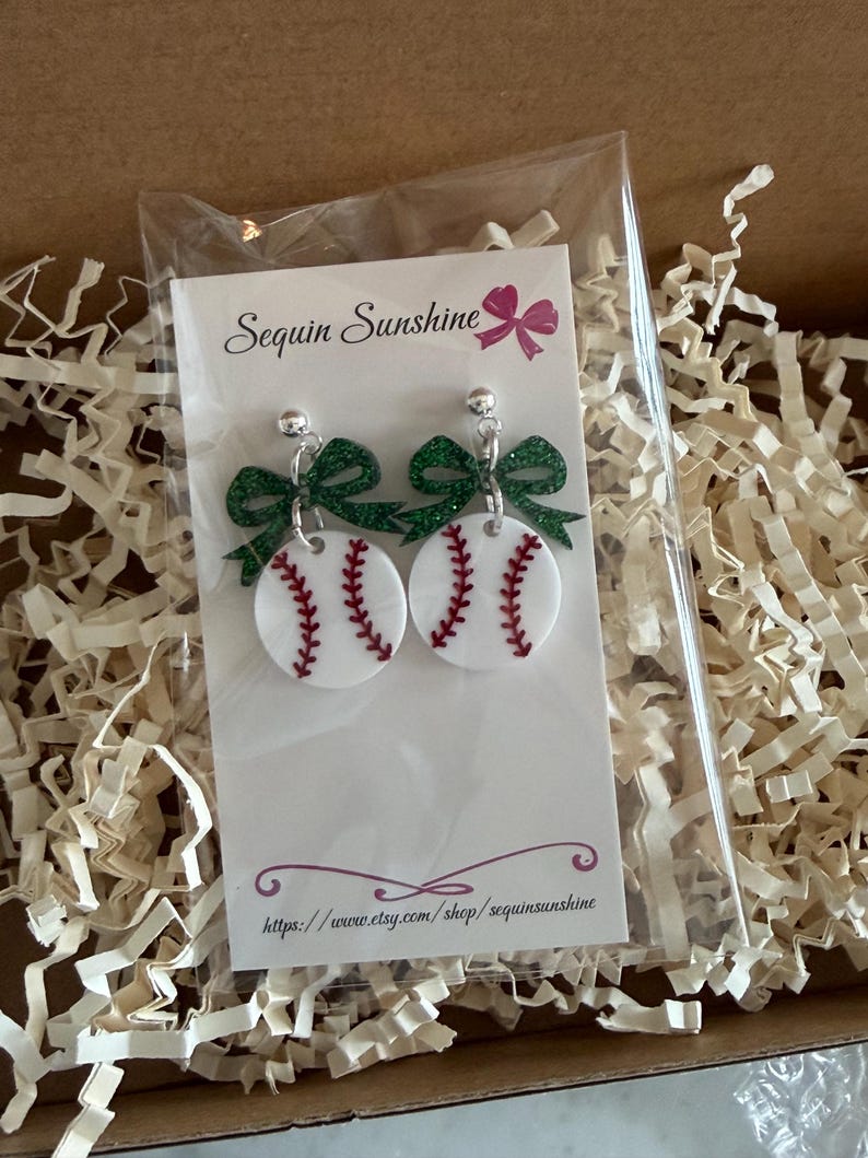 May include: Baseball-themed earrings with white baseballs featuring red stitching and green glitter bows. The earrings are displayed on a card with the shop name "Sequin Sunshine" and a website address, inside a cardboard box with paper filler.