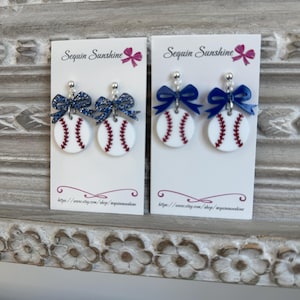 May include: Two pairs of baseball-themed earrings. Each earring features a white, circular design with red stitching, resembling a baseball. They are adorned with a blue bow and a silver-colored stud. The text "Sequin Sunshine" is visible.