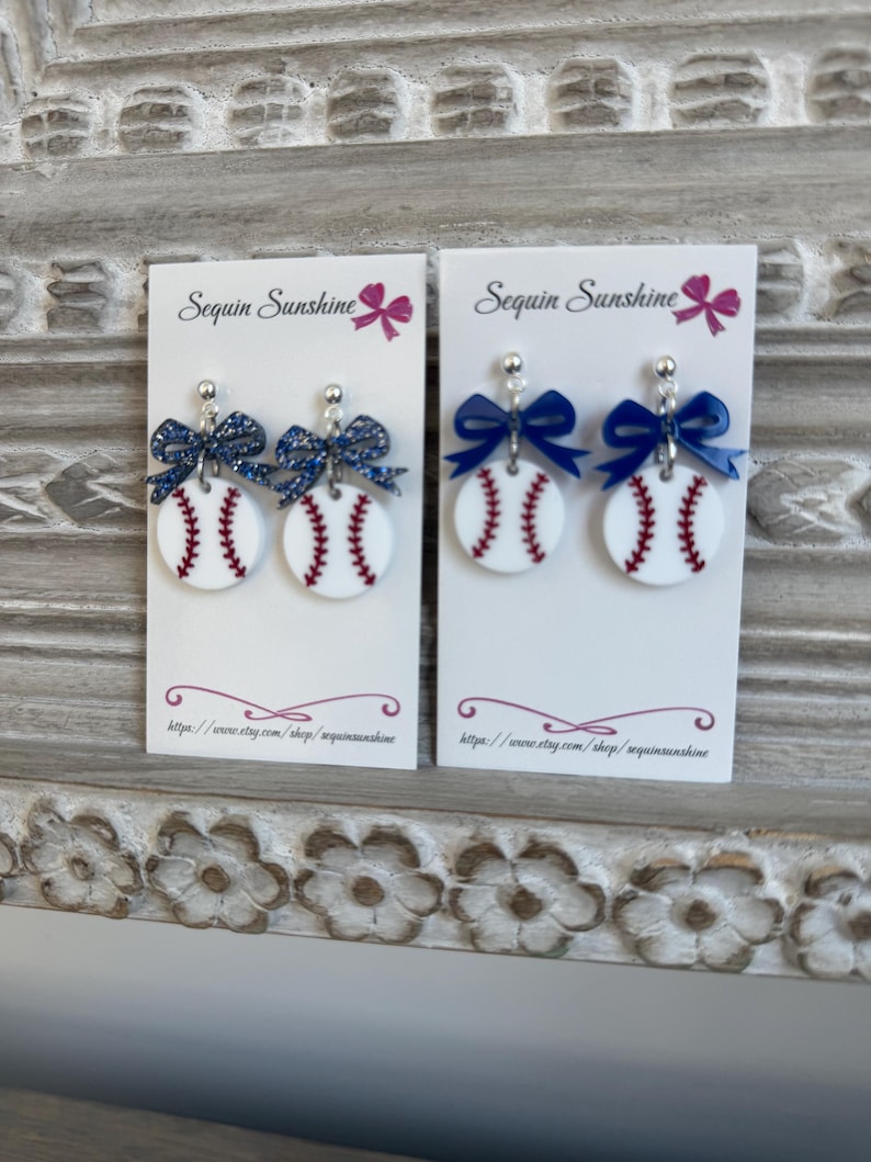 May include: Two pairs of baseball-themed earrings. Each earring features a white, circular design with red stitching, resembling a baseball. They are adorned with a blue bow and a silver-colored stud. The text "Sequin Sunshine" is visible.