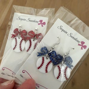 May include: Baseball-themed earrings with white baseballs featuring red stitching and a glittery bow. The earrings are packaged on a white card with the text "Sequin Sunshine" and a pink bow graphic. The earrings are on a light brown surface.