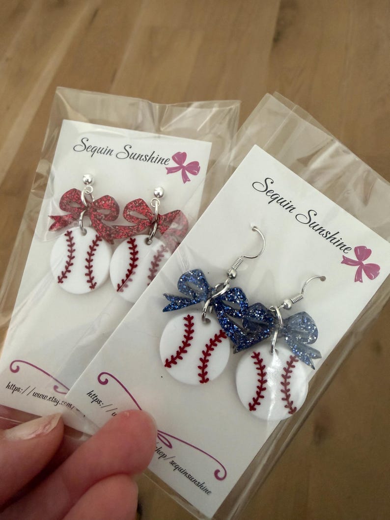 May include: Baseball-themed earrings with white baseballs featuring red stitching and a glittery bow. The earrings are packaged on a white card with the text "Sequin Sunshine" and a pink bow graphic. The earrings are on a light brown surface.