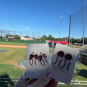 May include: Baseball-themed earrings. One pair features a baseball design with red stitching and a red bow. The other pair has a baseball design with red stitching and a red glitter top. The earrings are displayed against a baseball field background.