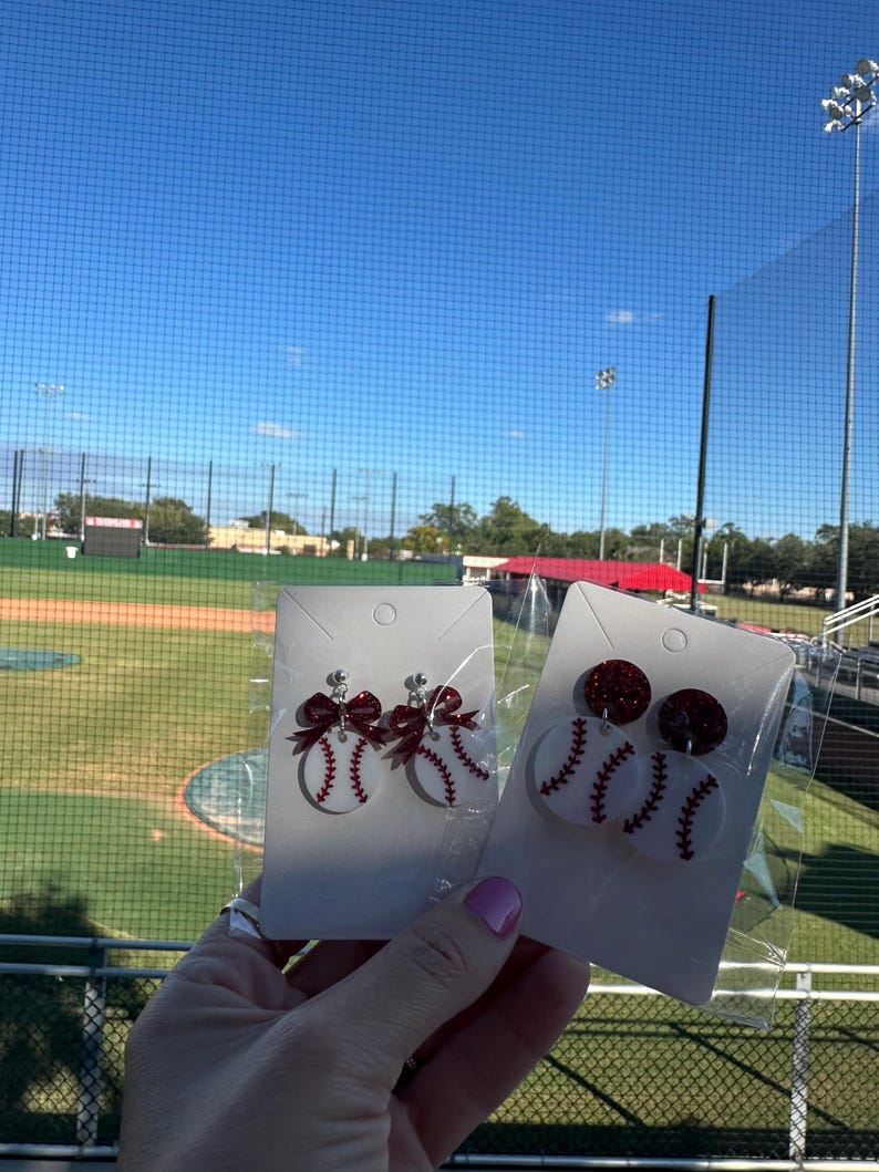 May include: Baseball-themed earrings. One pair features a baseball design with red stitching and a red bow. The other pair has a baseball design with red stitching and a red glitter top. The earrings are displayed against a baseball field background.