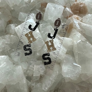 May include: A pair of dangle earrings featuring the letters "JHS" in black and gold glitter. Each earring has a football-shaped charm at the top. The earrings are displayed against a white, crystalline background.