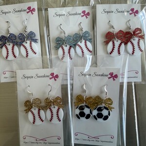 May include: Baseball and soccer ball themed earrings with glittery bows. The earrings are white with red stitching and a black and white soccer ball design. The earrings are on white cards with the text "Sequin Sunshine".