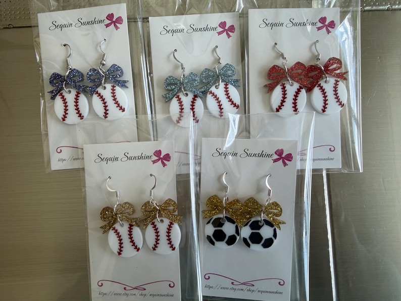 May include: Baseball and soccer ball themed earrings with glittery bows. The earrings are white with red stitching and a black and white soccer ball design. The earrings are on white cards with the text "Sequin Sunshine".