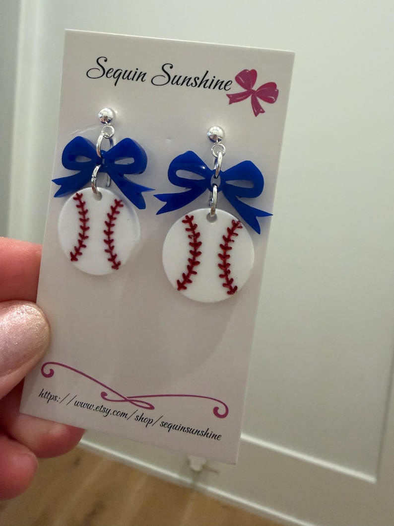 May include: Baseball-themed earrings featuring white circular pendants with red stitching, topped with a blue bow. The earrings are attached to a silver post. The card behind the earrings says "Sequin Sunshine" and has a website address.