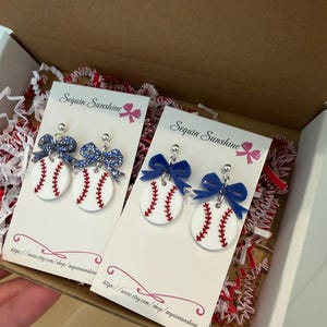May include: Baseball-themed earrings with white baseballs, red stitching, and blue bows. The earrings are displayed on white cards inside a cardboard box with red and white filler. The cards read "Sequin Sunshine".