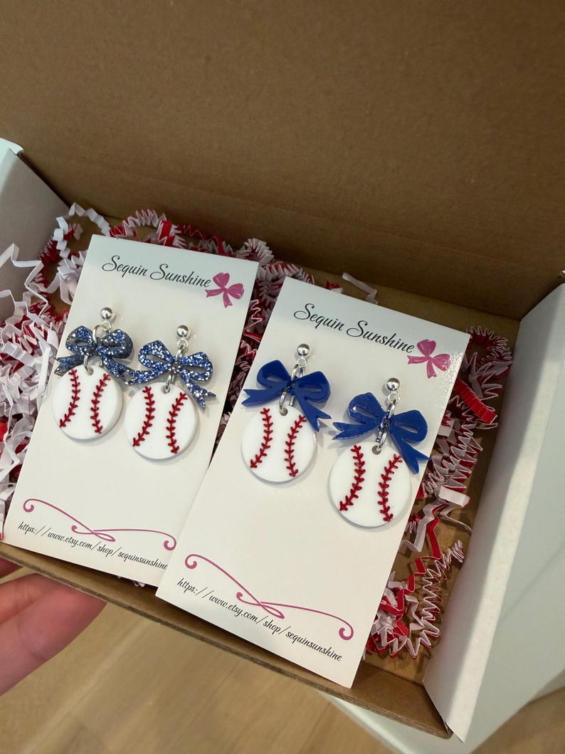 May include: Baseball-themed earrings with white baseballs, red stitching, and blue bows. The earrings are displayed on white cards inside a cardboard box with red and white filler. The cards read "Sequin Sunshine".
