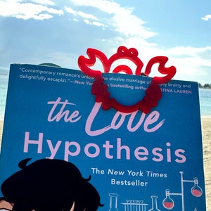 May include: A red plastic crab-shaped bookmark on a blue book with the title "The Love Hypothesis" and the text "The New York Times Bestseller".