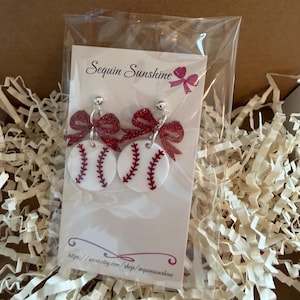 May include: Baseball-themed earrings featuring white circular designs with red stitching, resembling baseballs. Each earring has a red glitter bow at the top. The earrings are displayed on a white card with the text "Sequin Sunshine" and a website address.