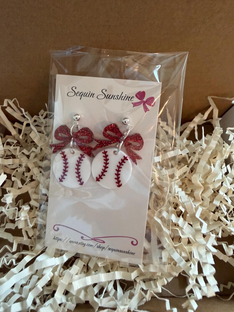 May include: Baseball-themed earrings featuring white circular designs with red stitching, resembling baseballs. Each earring has a red glitter bow at the top. The earrings are displayed on a white card with the text "Sequin Sunshine" and a website address.