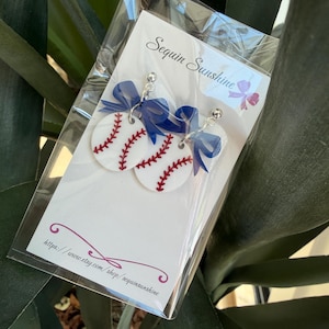 May include: Baseball-themed earrings featuring white circular pendants with red stitching, mimicking baseballs. Each earring has a blue bow and a silver-colored post. The earrings are packaged in a clear plastic bag with the text "Sequin Sunshine" and a website address.