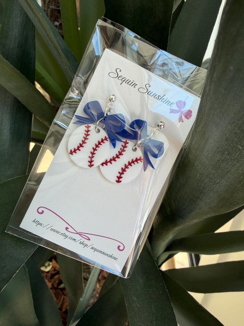 May include: Baseball-themed earrings featuring white circular pendants with red stitching, mimicking baseballs. Each earring has a blue bow and a silver-colored post. The earrings are packaged in a clear plastic bag with the text "Sequin Sunshine" and a website address.