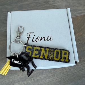 May include: A black and gold glitter keychain featuring the word SENIOR with 2027 integrated into the text, a graduation cap charm labeled Fiona, and a yellow tassel. The keychain is attached to a silver metal ring and rests on a white box.